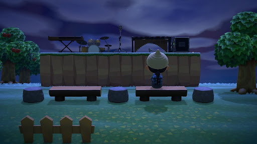 screenshot of Animal Crossing New Horizons where the player character is sitting on a wooden bench facing a stage. It is dark and cloudy out.