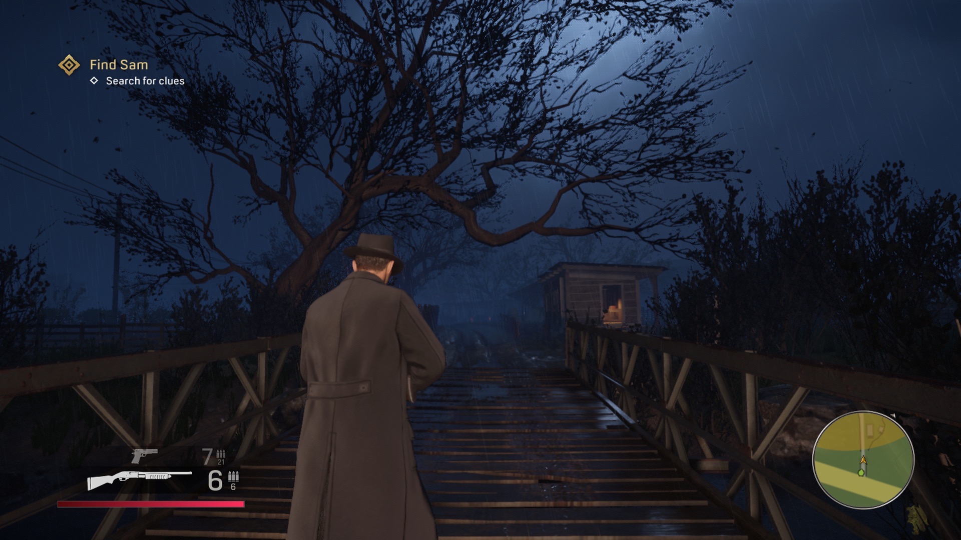 Gameplay screenshot in close third person perspective. Tommy's back is towards the camera as he walks across a bridge at night towards a tree and building on the other side. The shotgun is equipped and the mission is to search for clues