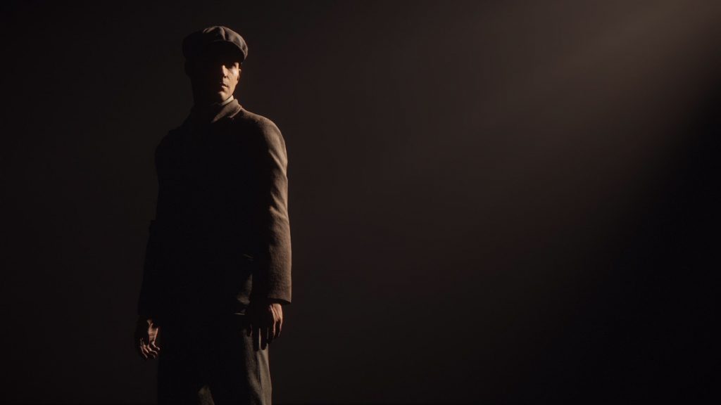 Tommy, a white man in a brown suit and cap standing against a dark background with moody lighting.