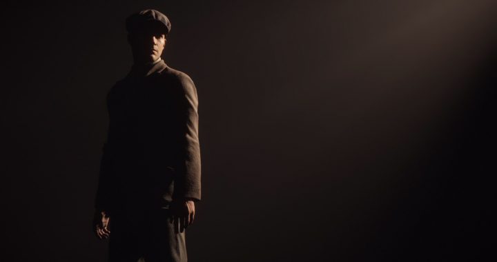 Tommy, a white man in a brown suit and cap standing against a dark background with moody lighting.