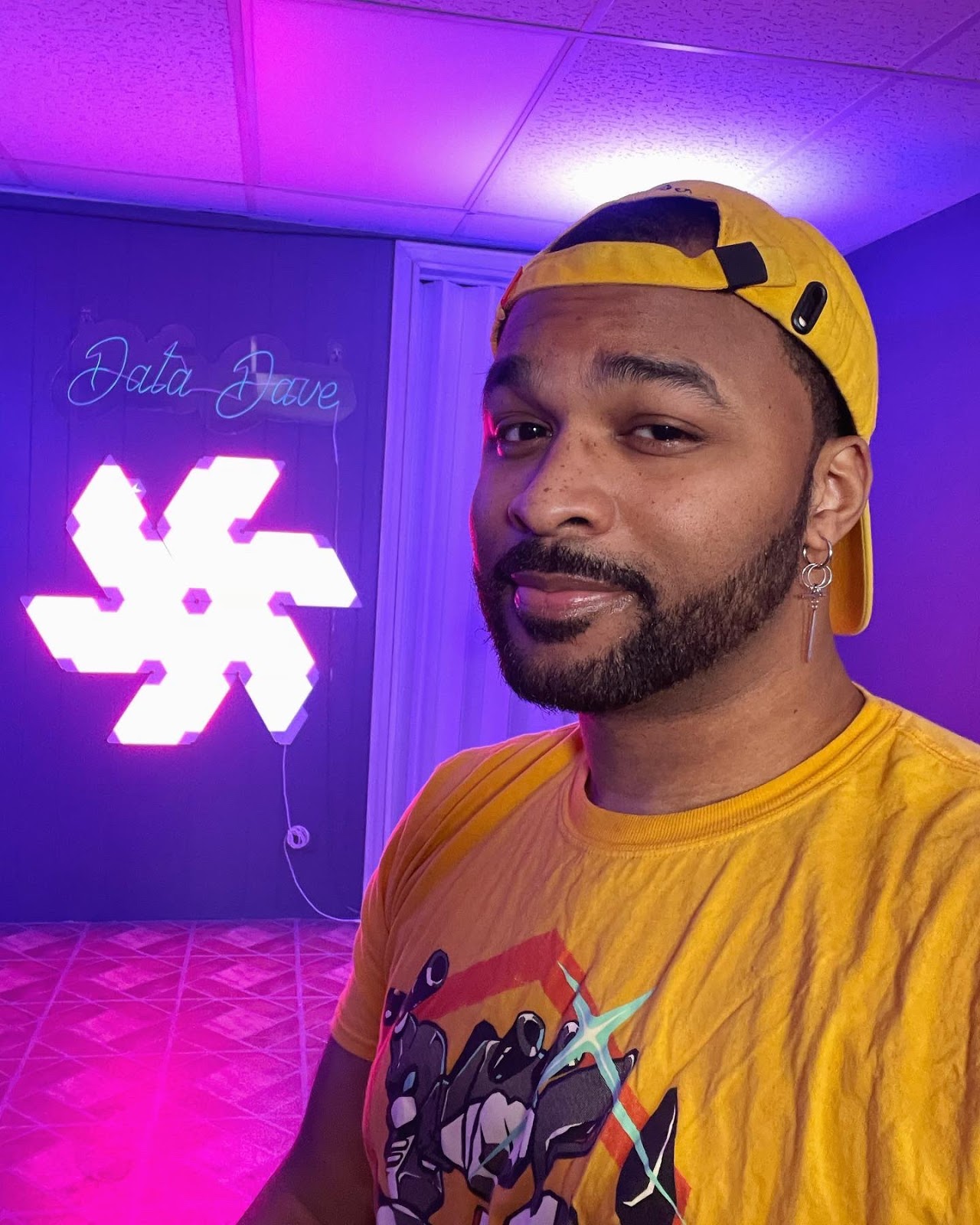 DataDave, a Black man in his 20'-30's wearing a yellow shirt and yellow backwards ball cap. The room behind him is glowing with purple light