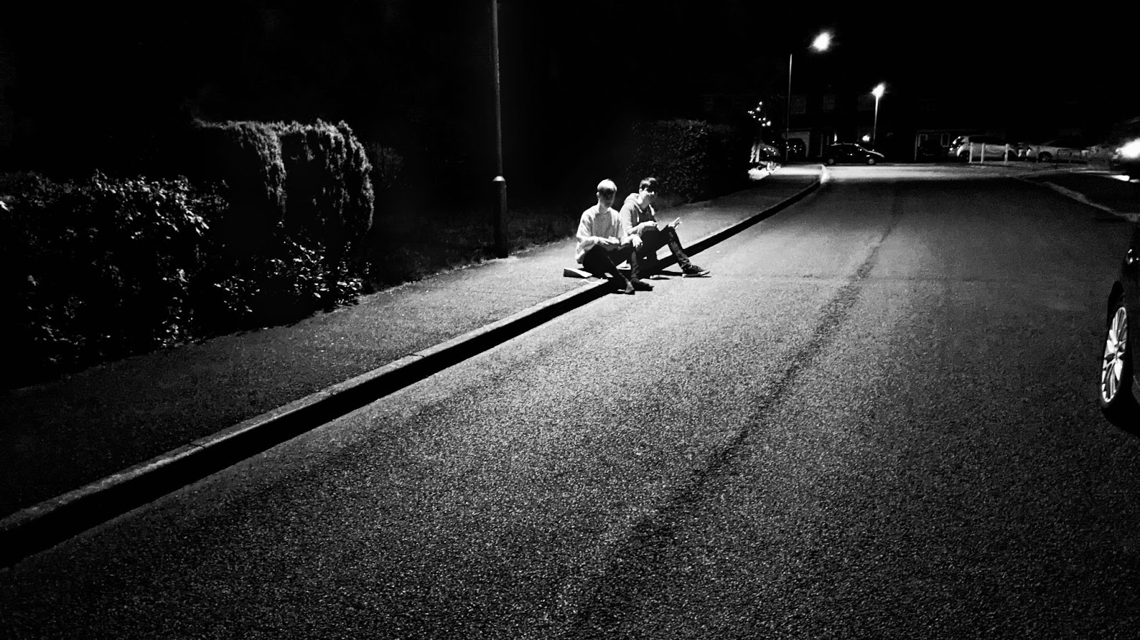 A black and white photo of two boys sitting on a curb under a street light
