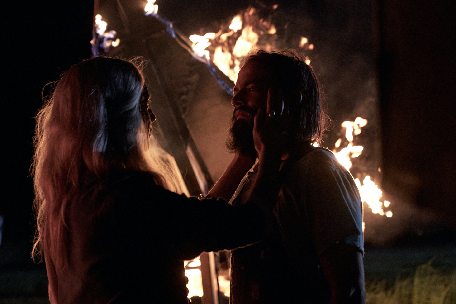 Caitlin McRill Fitzgerald holding Daniel Zovatto's face as a fire burns behind them in the darkness