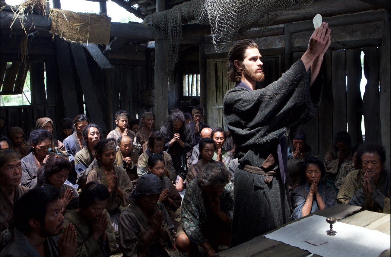 Andrew Garfield holding up a stone while Japanese people pray behind him in Silence